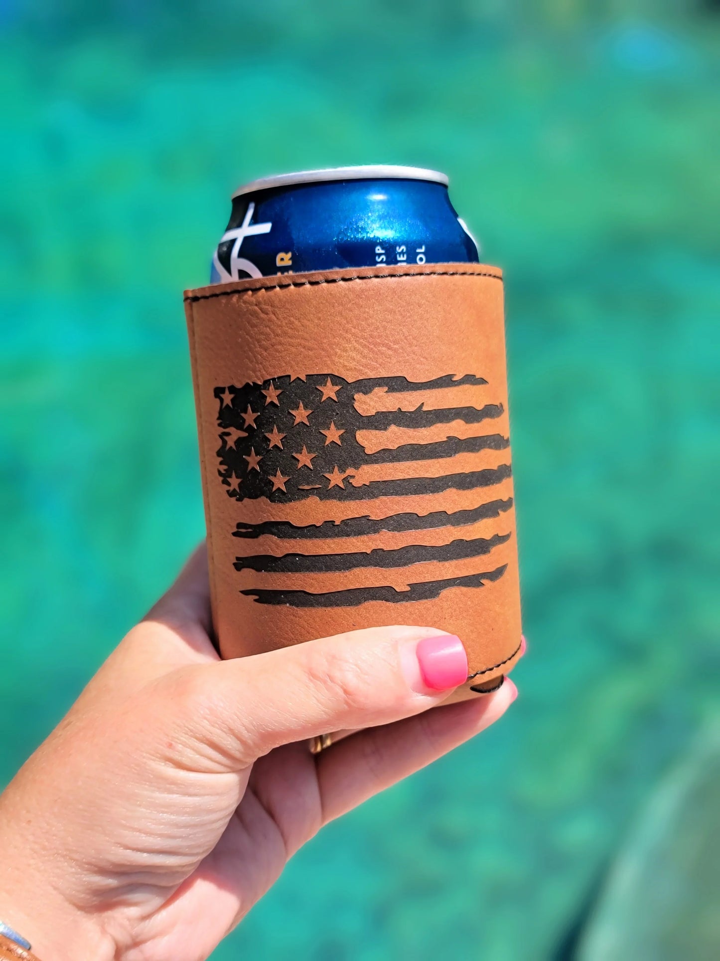 Hand holding a can in a brown leather koozie with engraved American flag design by a pool.
