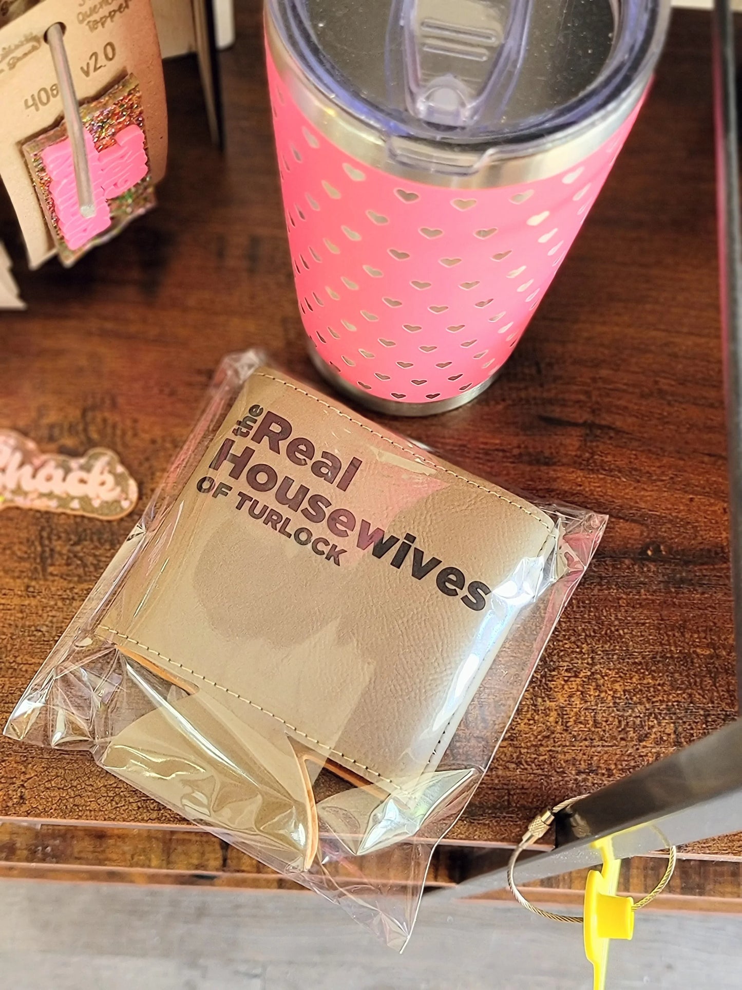 Pink tumbler with heart pattern and a beige wallet labeled 'Real Housewives of Turlock' on wooden shelf