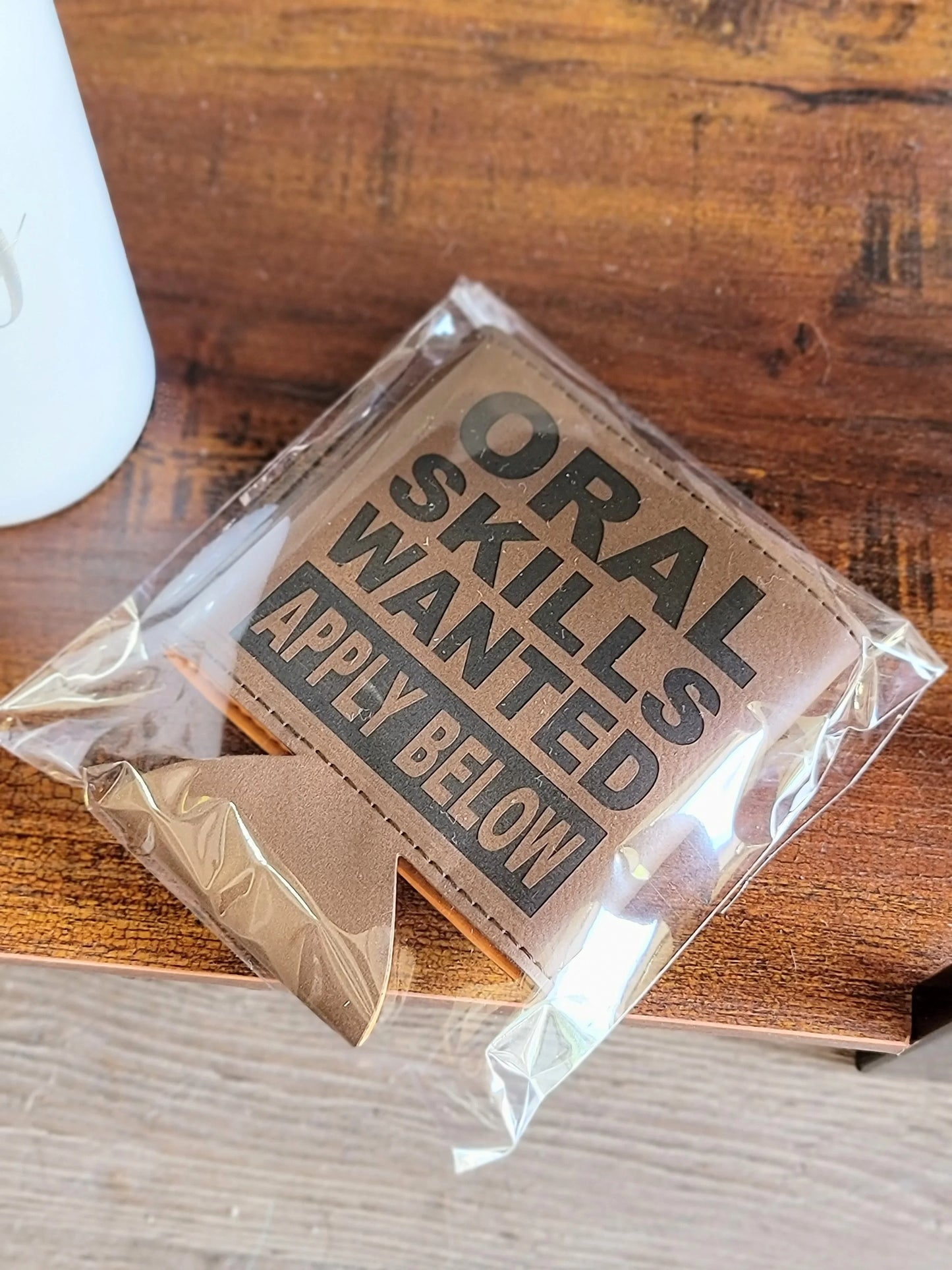 Brown leather drink koozie with humorous engraved text, packaged on a wooden table